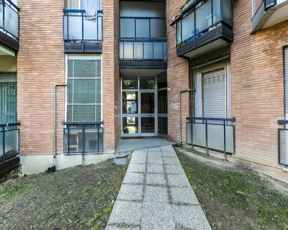Exterior view of an apartment building entrance with a paved walkway, ground-level balconies and glass doors leading into the lobby.