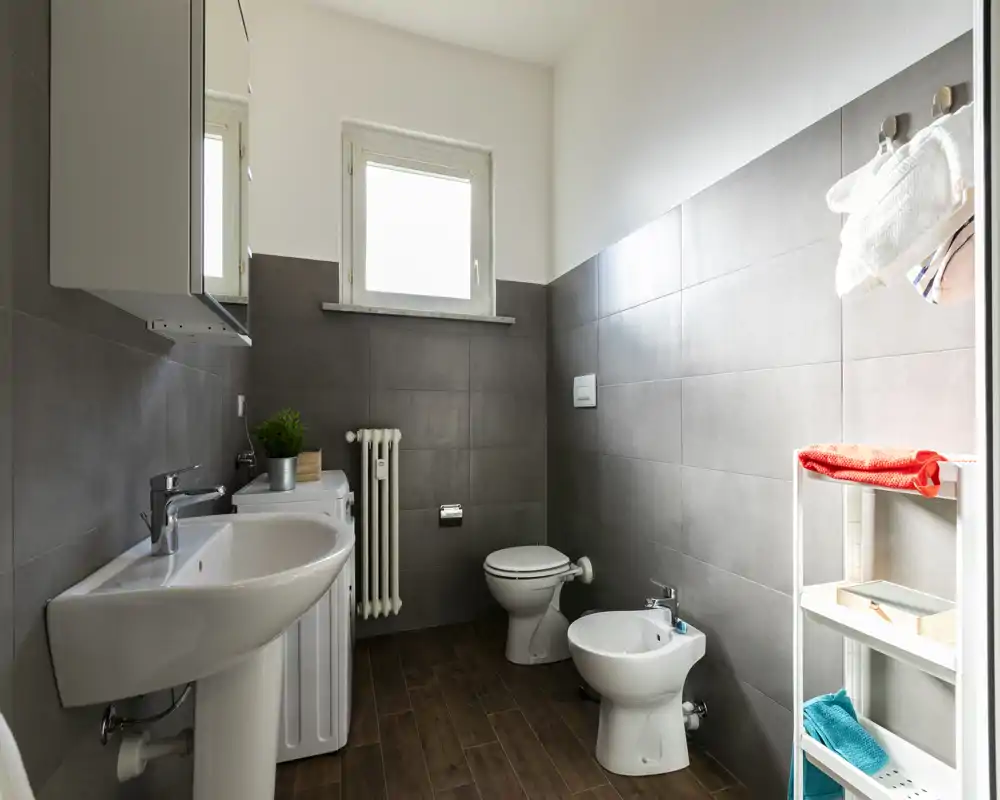 Bright, modern bathroom with wall-mounted sink, toilet and bidet, tiled walls and wood-effect floor; neat shelving with towels and a small window providing natural light.