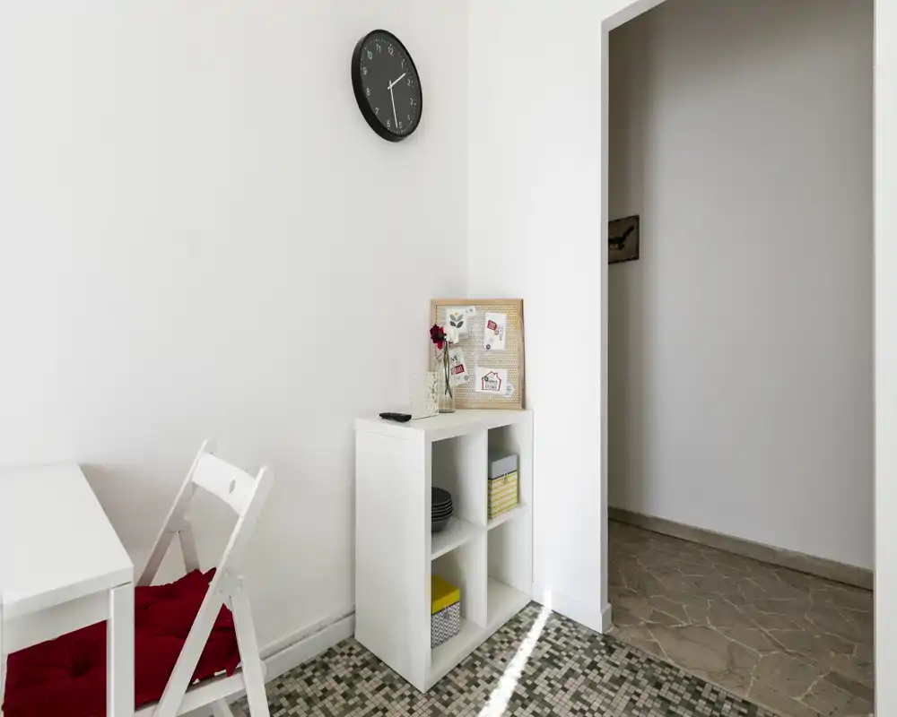 Bright, minimal living area corner with a white shelving unit, folding chair with red cushion, and a small desk — simple, well-lit space suitable for short stays.