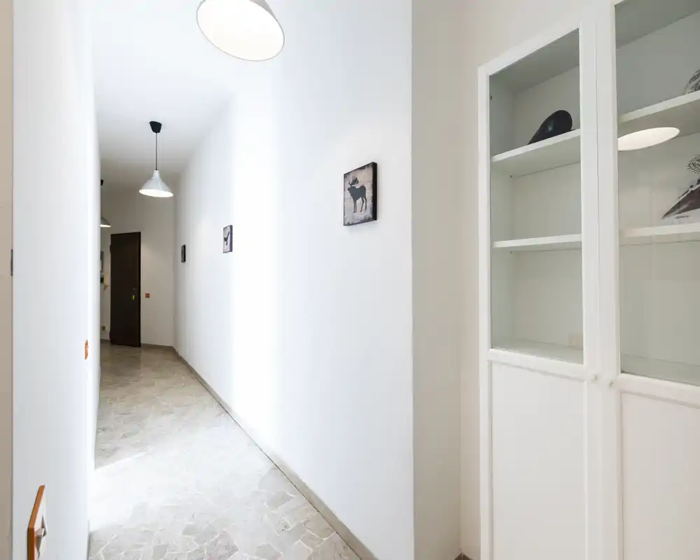 Bright, minimal hallway/entrance with tiled floor, pendant lighting and a built-in glass-front cabinet; clean white walls and small wall art create a neat first impression.