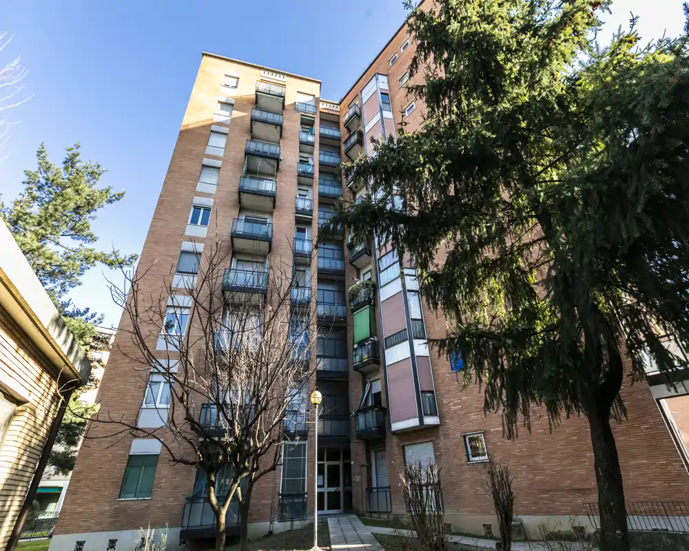 Exterior view of a multi-story brick apartment building with balconies, a small courtyard and mature trees under bright daylight.