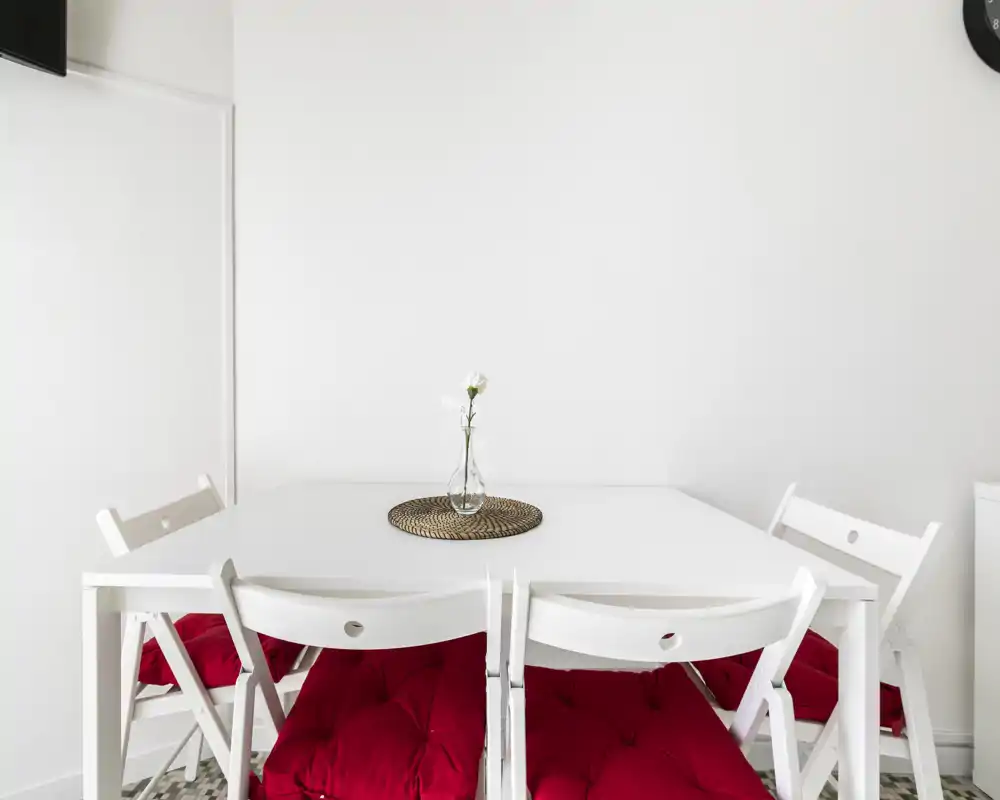 Bright minimalist dining area with a white table and four white folding chairs topped with red seat cushions; a small glass vase with a single flower creates a simple, elegant centerpiece.