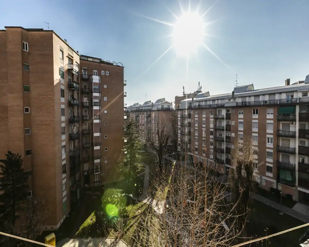Sunny external view of a residential courtyard framed by multi-storey brick apartment buildings, with trees and a walkway below — ideal for showcasing a balcony or terrace outlook.