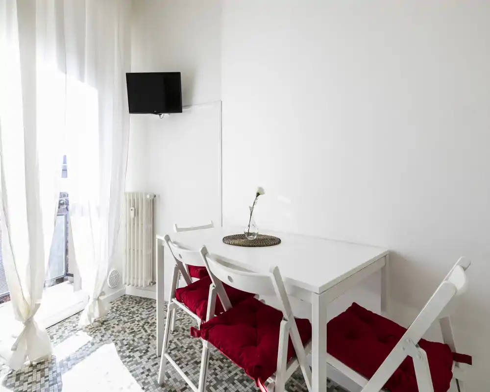 Compact dining area with a white table and folding chairs topped with red seat cushions, lit by natural light from a balcony and styled with a single flower centerpiece.