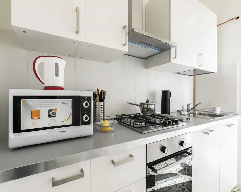 Modern, compact kitchen with stainless worktop, gas hob, built-in oven and countertop appliances. Bright, clean design with white cabinetry and practical utensils.