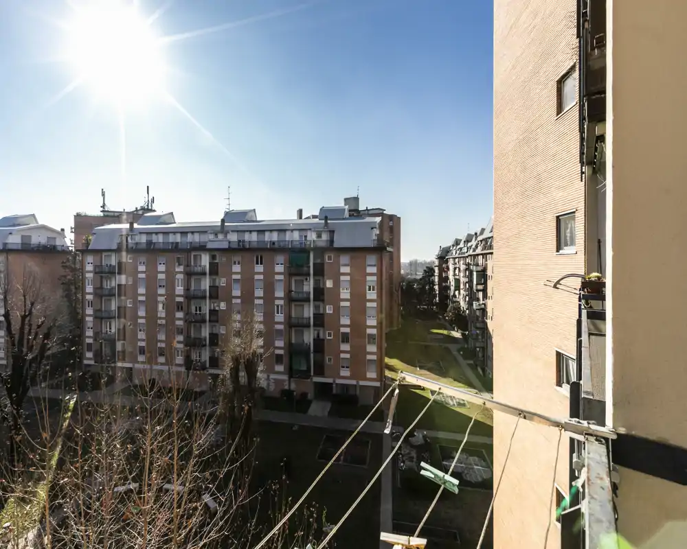 Sunny view from a balcony overlooking neighboring apartment buildings and a green courtyard; clear blue sky and bright natural light.