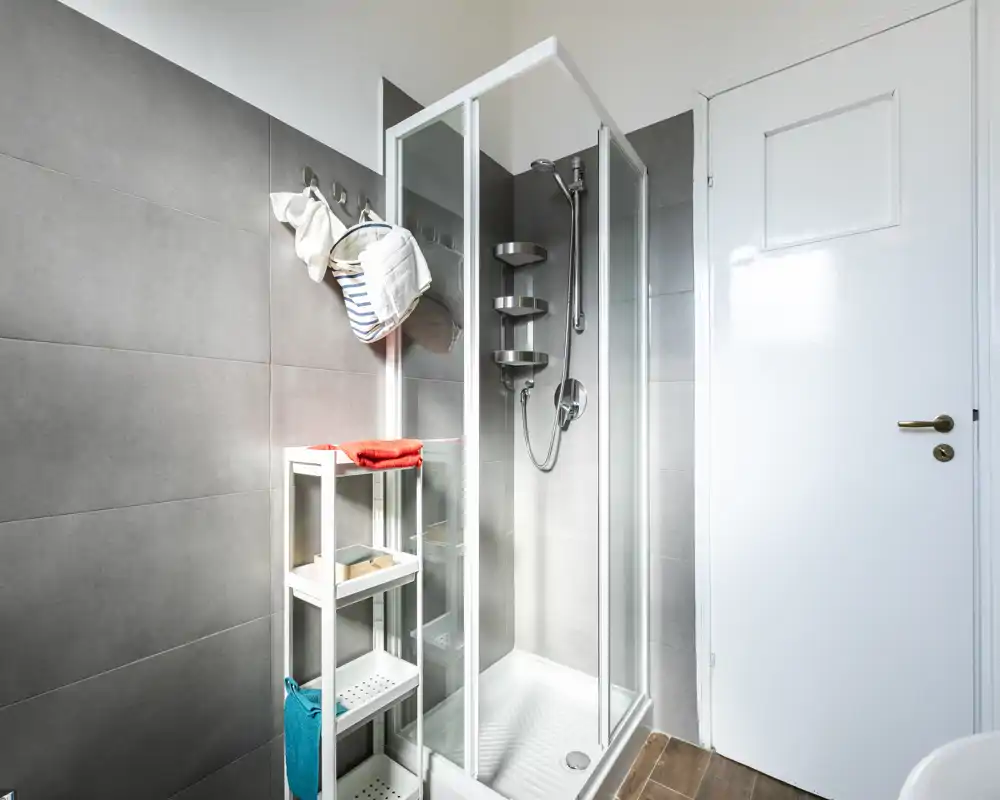 Modern, compact bathroom with a glass shower cabin, wall-mounted shower head, and tidy shelving for toiletries and towels. Bright neutral tiles and wooden-effect flooring create a clean, contemporary look.