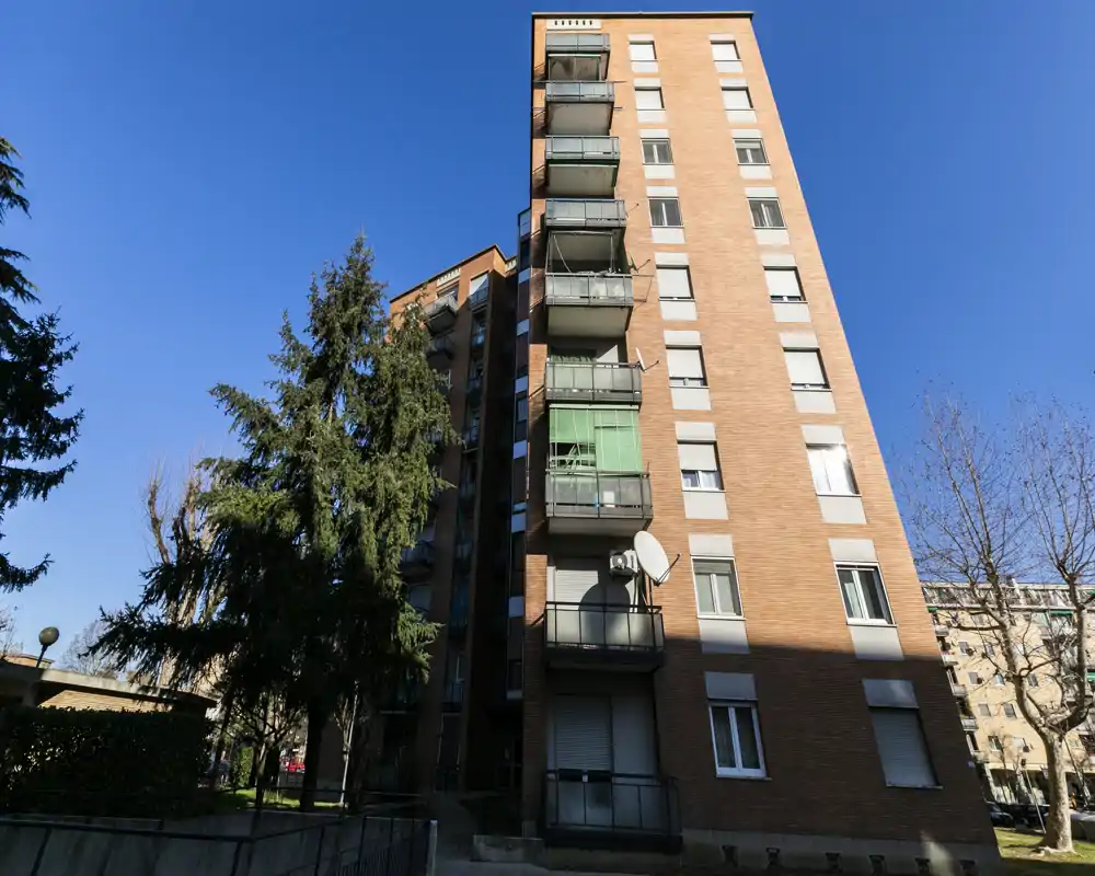 Exterior view of a multi-storey brick apartment building with balconies, satellite dish and surrounding trees under a clear blue sky.