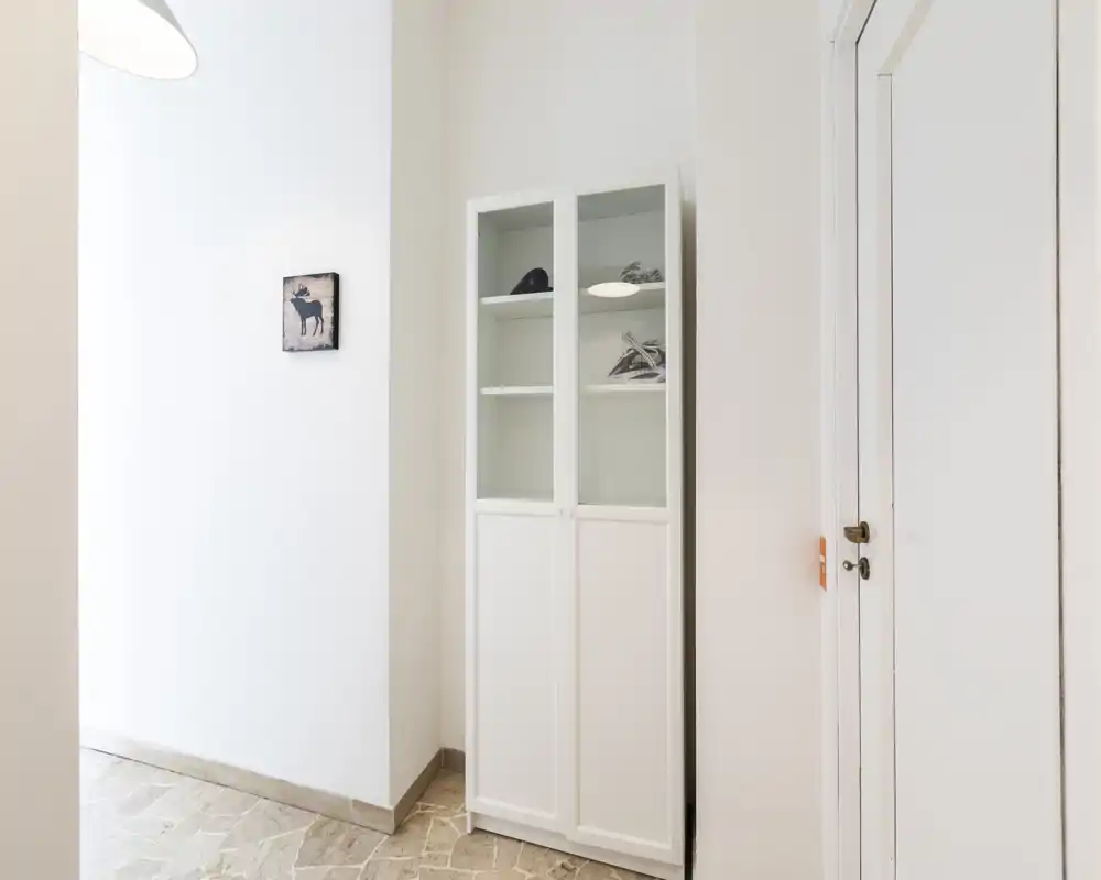 A bright, minimal entrance/hallway with a tall white storage cabinet, pale tiled floor and a simple wall decoration. Clean lines and neutral tones create a welcoming arrival space.