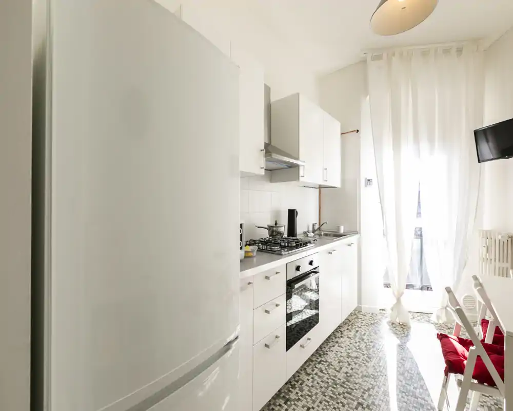 Bright modern galley kitchen with white cabinets, built-in oven and gas hob, fridge at the foreground and a sunlit balcony at the end — compact and inviting for short stays.