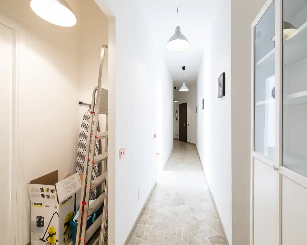 Bright, narrow corridor/entrance with stone-effect floor tiles, pendant lights and a built-in storage cabinet; currently contains some household items and a ladder in a small alcove.