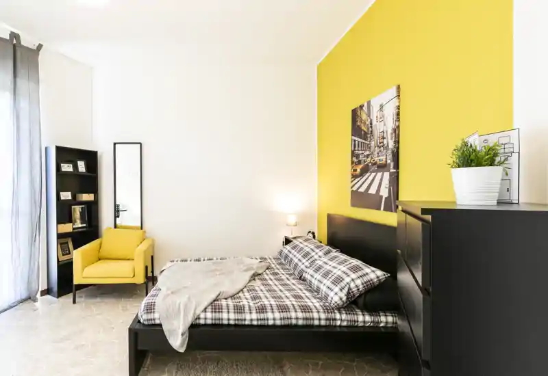 Chambre lumineuse et moderne avec un lit double en parure à carreaux, un mur d'accent jaune et un fauteuil confortable près de la fenêtre.