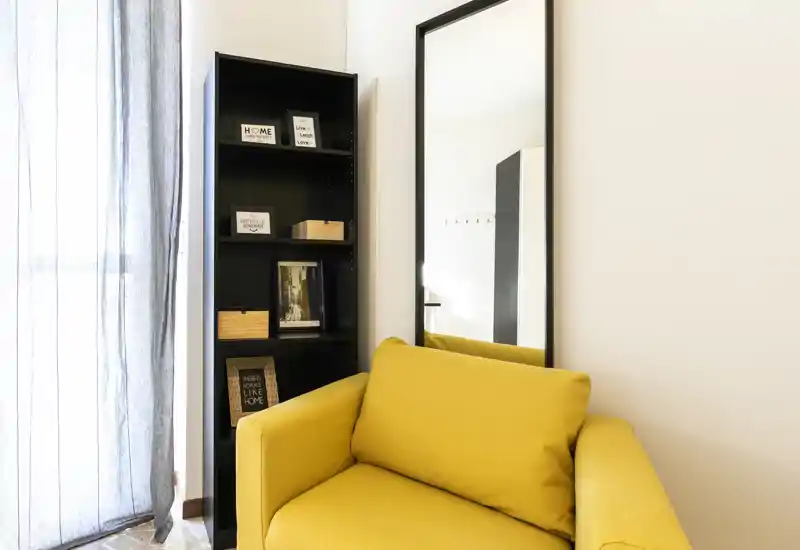 Angolo moderno e luminoso con una comoda poltrona gialla, uno specchio alto e una sottile libreria nera: perfetto per un accogliente angolo living con luce naturale.