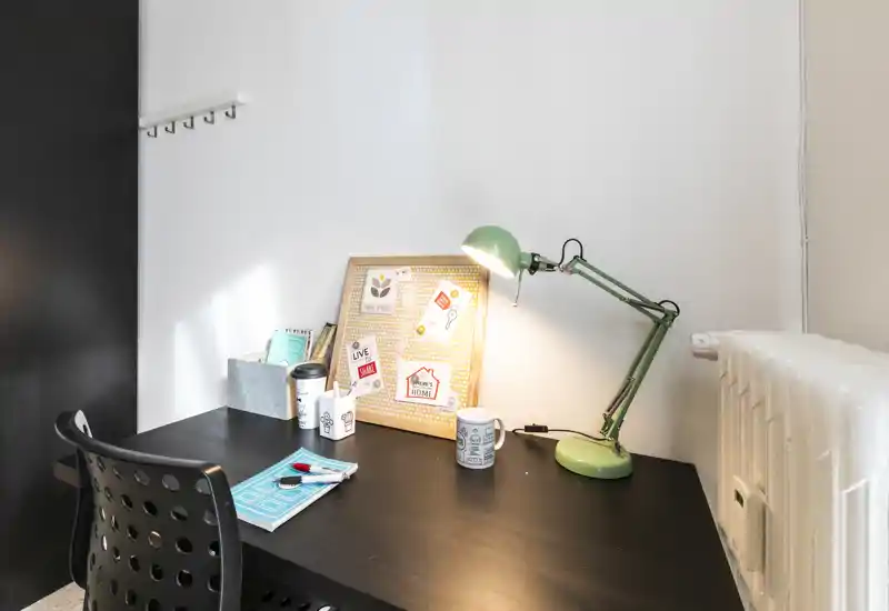 Spazio di lavoro luminoso e ordinato con scrivania nera, lampada da tavolo verde regolabile, bacheca e sedia forata — perfetto per leggere o lavorare da casa.