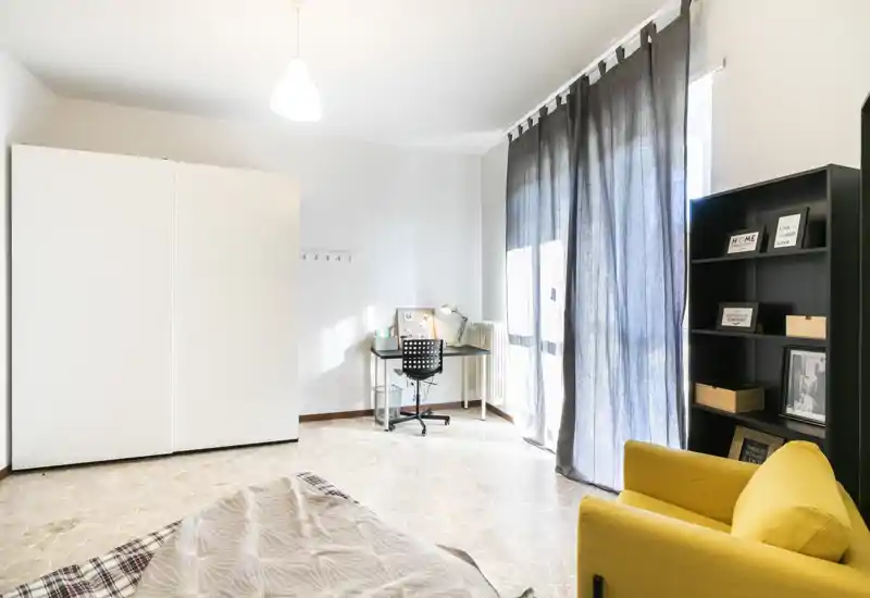Camera da letto moderna e luminosa con letto in primo piano, scrivania vicino alla finestra, grande armadio e una comoda poltrona gialla—ideale per un soggiorno confortevole.