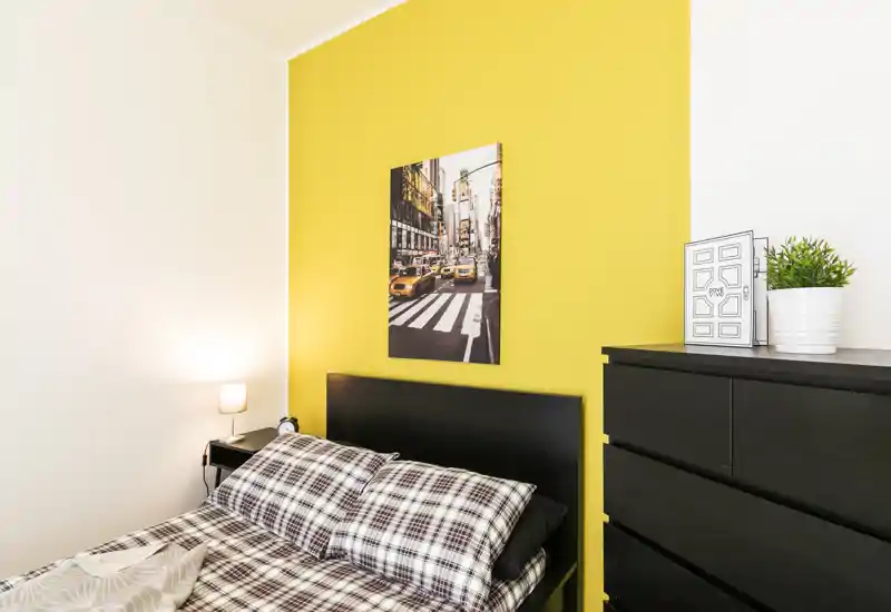Chambre moderne et lumineuse avec tête de lit noire sur un mur jaune, linge à carreaux, lampe de chevet et commode avec plante décorative — propre et bien mise en scène pour une annonce.