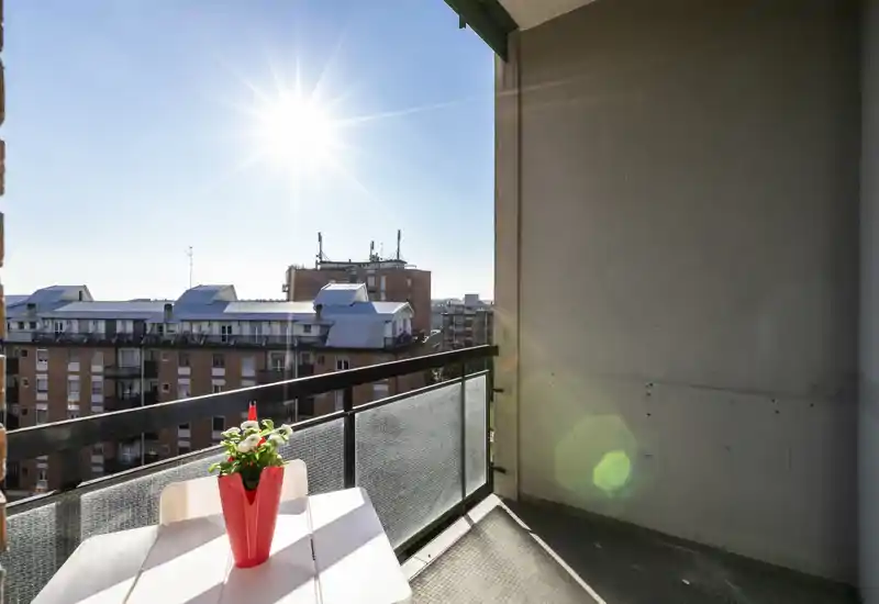 Balcone soleggiato con un piccolo tavolo e un vaso rosso decorativo, vista sulla città e ringhiera — perfetto per il caffè mattutino.