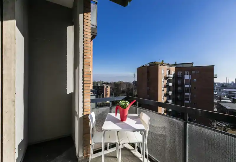 Balcone soleggiato con tavolino e due sedie, un vaso decorativo a forma di annaffiatoio rosso e vista sulla città — perfetto per il caffè mattutino.