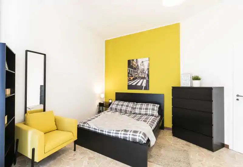Chambre lumineuse et moderne avec lit double devant un mur jaune vif, fauteuil jaune confortable, grande commode et lampe de chevet — idéale pour les annonces locatives.