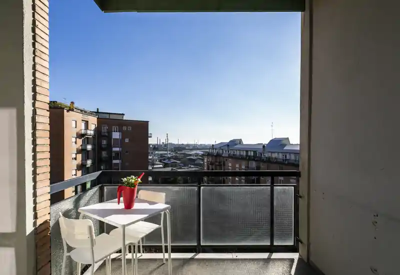 Balcone soleggiato con un piccolo tavolino bistro bianco, due sedie e un vaso rosso decorativo, con vista sui palazzi in mattoni e un cielo azzurro.