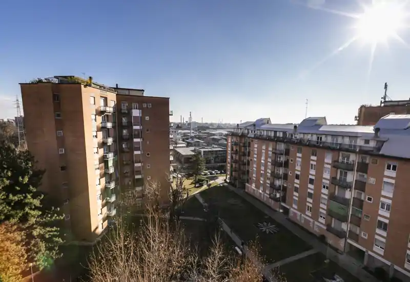 Vista luminosa del cortile urbano da un piano alto, con edifici residenziali, percorsi paesaggistici e skyline cittadino sullo sfondo in pieno giorno — perfetta per mostrare l’esterno e i dintorni dell’edificio.