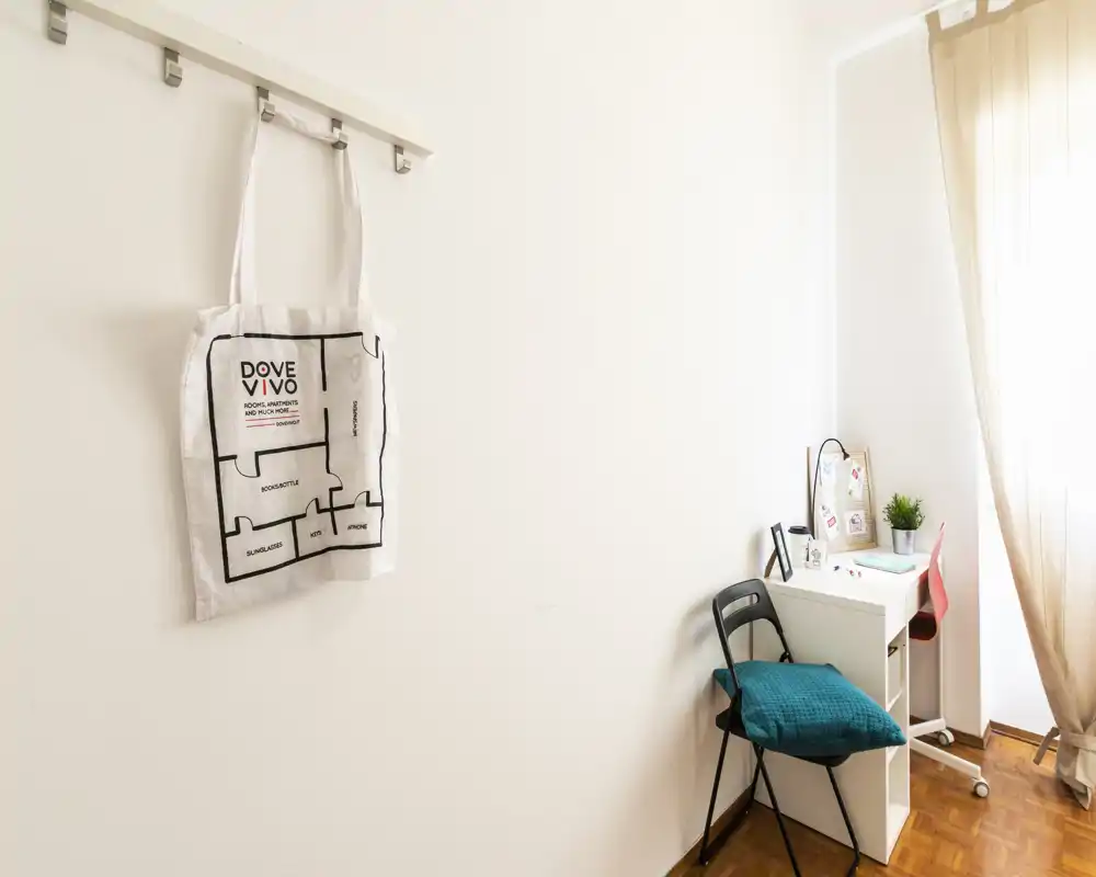 Petit coin lumineux avec un bureau compact, une chaise et une grande fenêtre voilée ; rares indices de chambre à coucher et déco minimaliste.