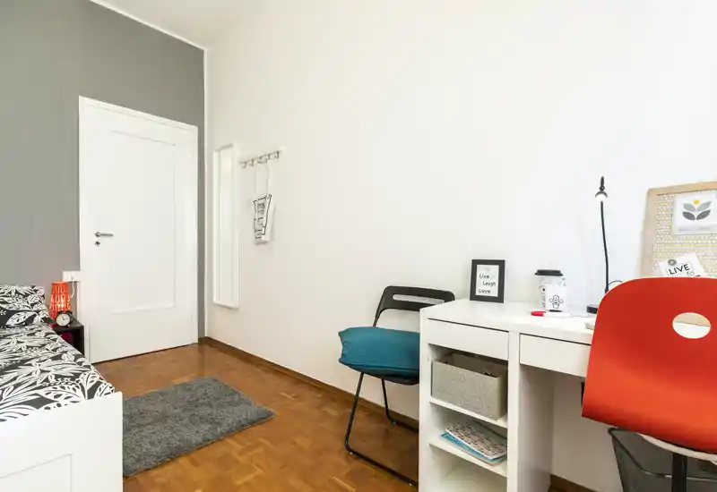 Chambre lumineuse et soignée avec un lit simple à motifs, un bureau et une chaise—idéale pour étudiant ou personne seule. La pièce est bien éclairée et décorée avec coussins et lampe de bureau.