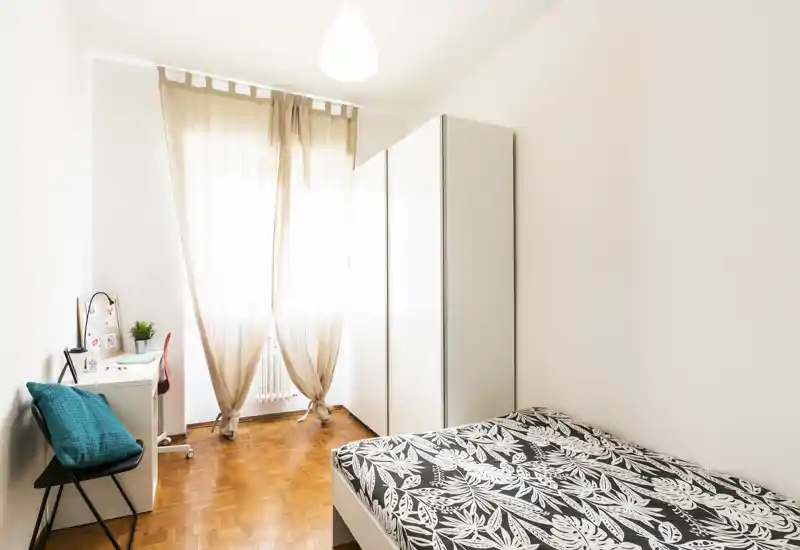 Chambre lumineuse et minimaliste avec un lit simple à motif, une grande armoire et un petit bureau près d'une fenêtre laissant entrer une belle lumière naturelle.