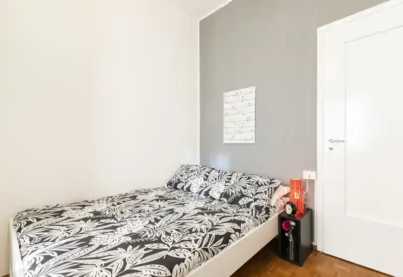 Chambre lumineuse et soignée avec un lit double habillé d'une parure à motif, une lampe de chevet et quelques objets décoratifs pour une atmosphère accueillante.