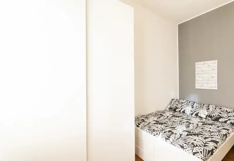 Chambre compacte avec lit double habillé de literie noire et blanche à motif botanique, mur d'accent neutre et mobilier minimaliste. Lumière naturelle abondante et placard discret pour un espace de nuit moderne et soigné.