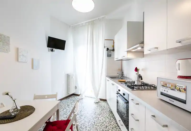 Cucina moderna e luminosa con mobili bianchi, piano cottura a gas, forno incassato e un piccolo tavolo da pranzo vicino a una grande finestra/porta che lascia entrare molta luce naturale.