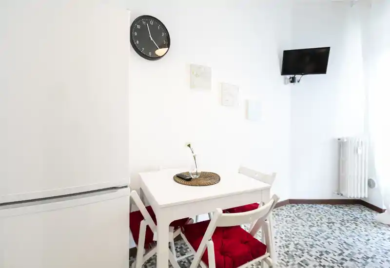 Piccola zona pranzo con tavolo bianco e quattro sedie con cuscini rossi, luce naturale intensa e decorazioni minimali per un aspetto moderno e pulito.