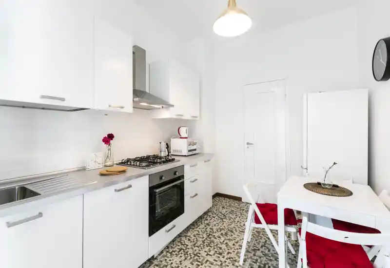 Cucina compatta e moderna con mobili bianchi, piano cottura a gas, forno incassato e piccolo tavolo da pranzo con cuscini rossi — luminosa, pulita e ben attrezzata per soggiorni brevi.