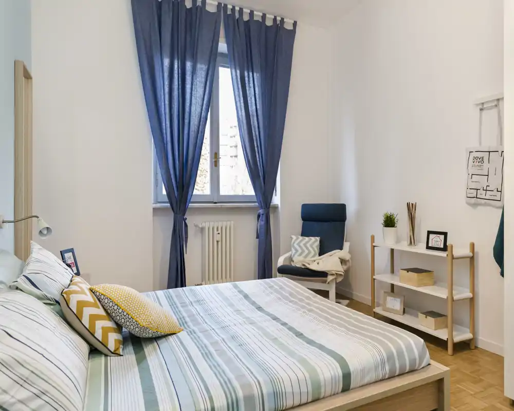 Dormitorio luminoso y bien presentado con la cama en primer plano, cortinas azules y un sillón de lectura junto a la ventana. Textiles suaves y cojines coordinados crean un ambiente moderno y relajante perfecto para anuncios.