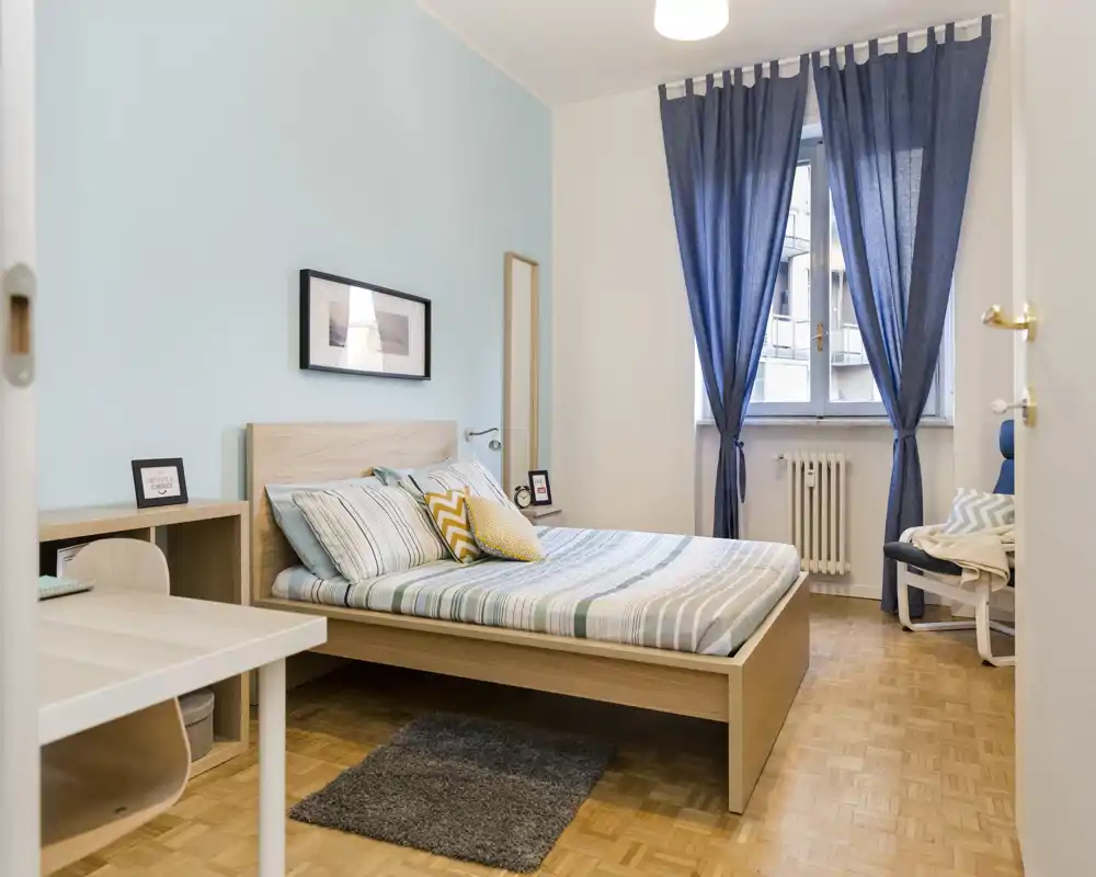 Dormitorio luminoso y bien presentado con cama doble, ropa de cama a rayas y cojines decorativos, un escritorio y una ventana con cortinas oscuras que dejan pasar la luz natural.