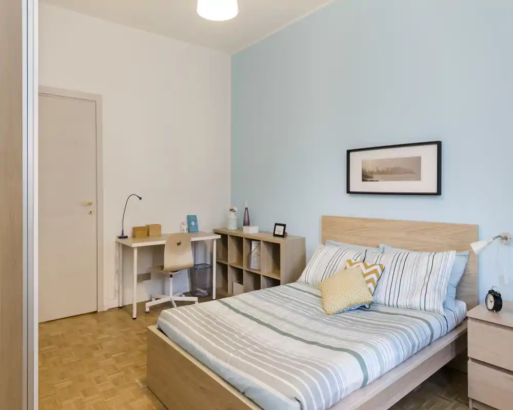 Dormitorio moderno y luminoso con cama doble, ropa de cama a rayas y cabecero de madera clara. La habitación incluye un escritorio compacto, estanterías y mesita de noche — limpia, minimalista y acogedora.