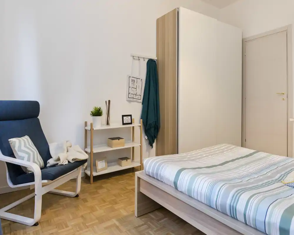 Dormitorio moderno y luminoso con cama doble visible en estructura de madera clara y ropa de cama a rayas, sillón cómodo y armario — estilo limpio y minimalista para anuncios de alquiler.
