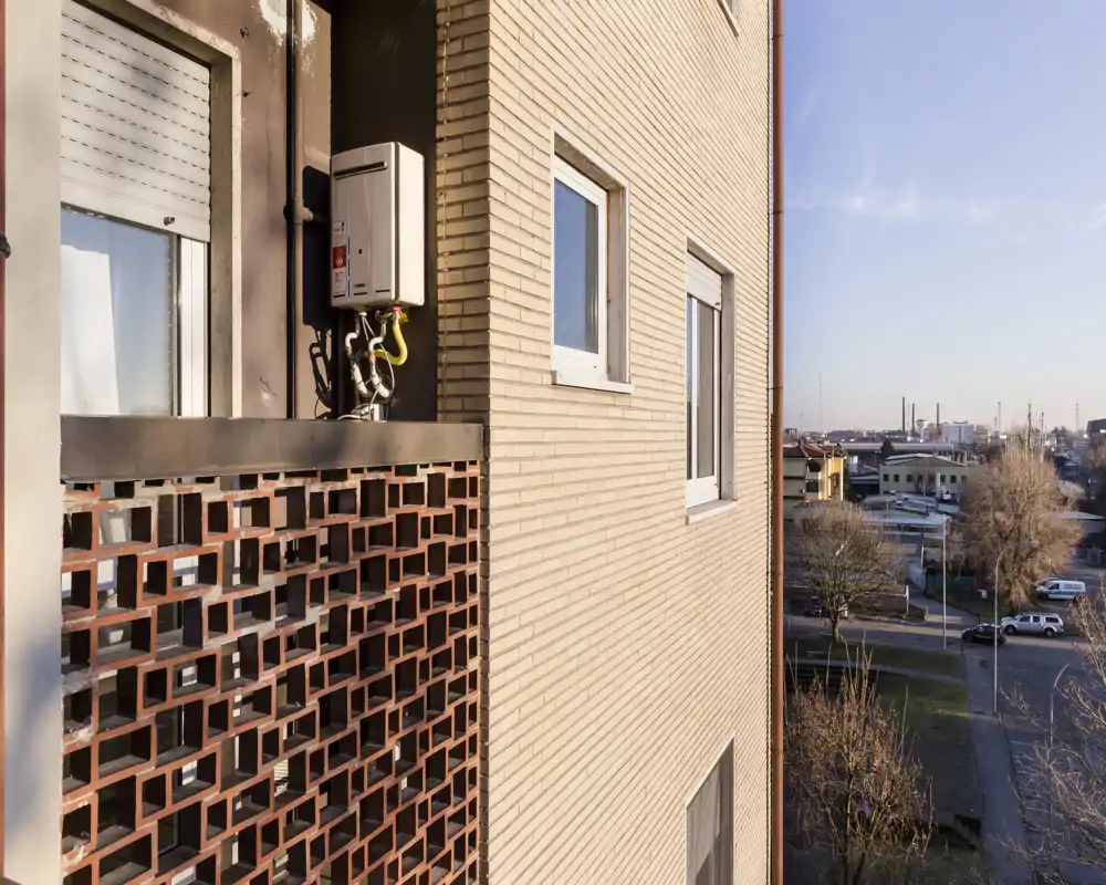 Pequeño balcón exterior con antepecho de ladrillo perforado, vista a una calle residencial y zona industrial en la distancia; buena luz natural al atardecer.