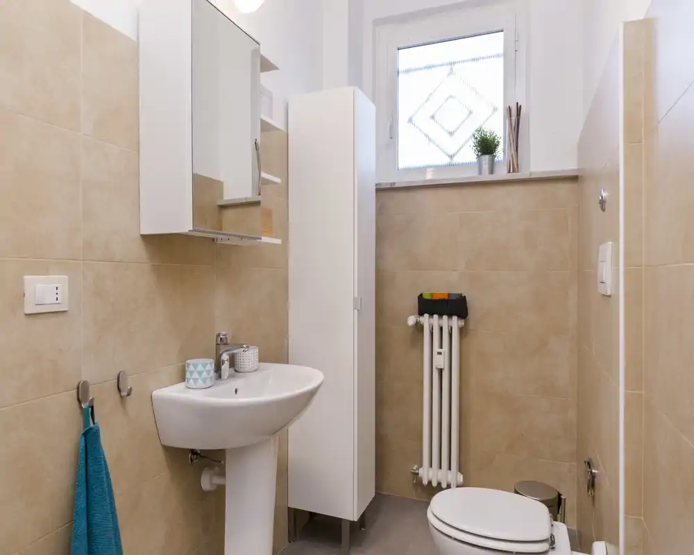 Baño pequeño y moderno, limpio, con lavabo de pedestal, mueble espejo, columna de almacenamiento, radiador y WC; azulejos beige neutros y luz natural desde una ventana translúcida.