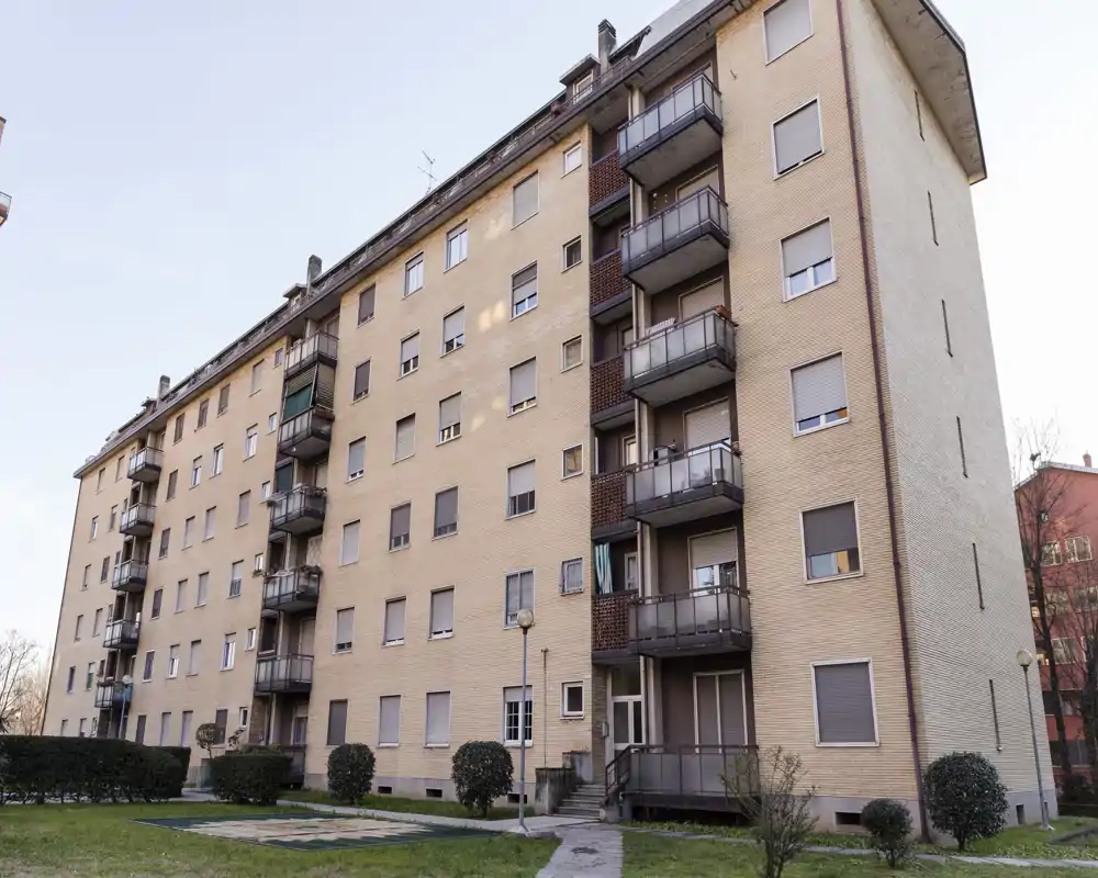 Vista exterior de un edificio residencial de varios pisos con balcones, zonas verdes en planta baja y un camino pavimentado hacia la entrada. Ideal para anuncios que destaquen la fachada, el acceso a los balcones y las zonas exteriores comunes.