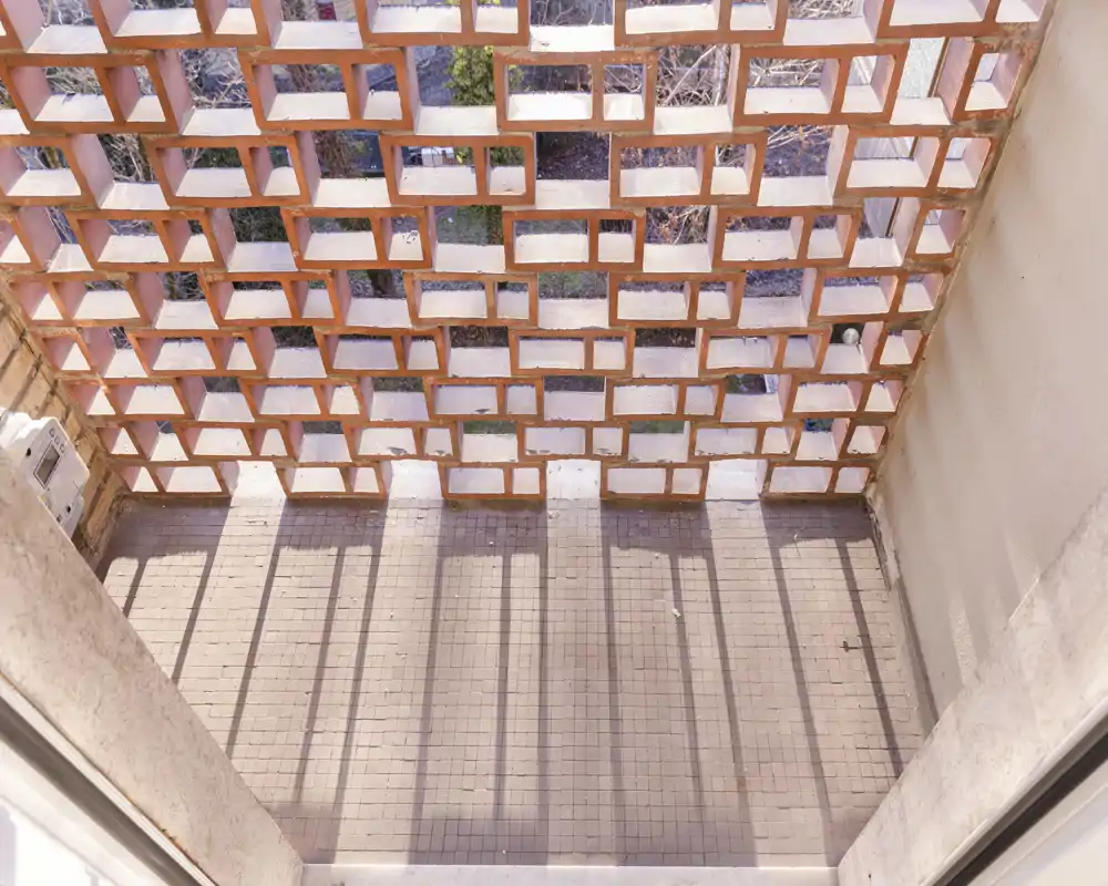 Zona de balcón/terraza exterior con cerramiento de ladrillo calado que proyecta sombras en rejilla sobre el suelo de baldosas. Ideal para una pequeña zona de estar o plantas, con mucha luz.