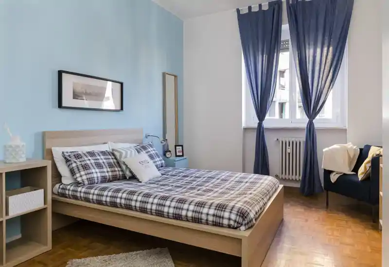 Chambre lumineuse et mise en scène, lit double avec linge à carreaux assorti et coussins décoratifs ; la grande fenêtre et le mur bleu apportent une atmosphère chaleureuse.