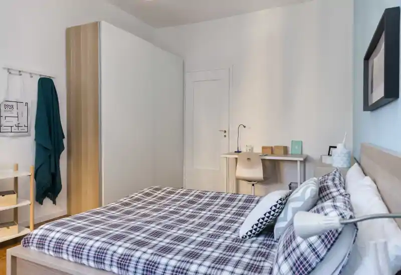 Camera da letto luminosa e ben arredata con un grande letto con copriletto a quadri, lampada da comodino e una piccola scrivania: perfetta per annunci in affitto che puntano su comfort e funzionalità.