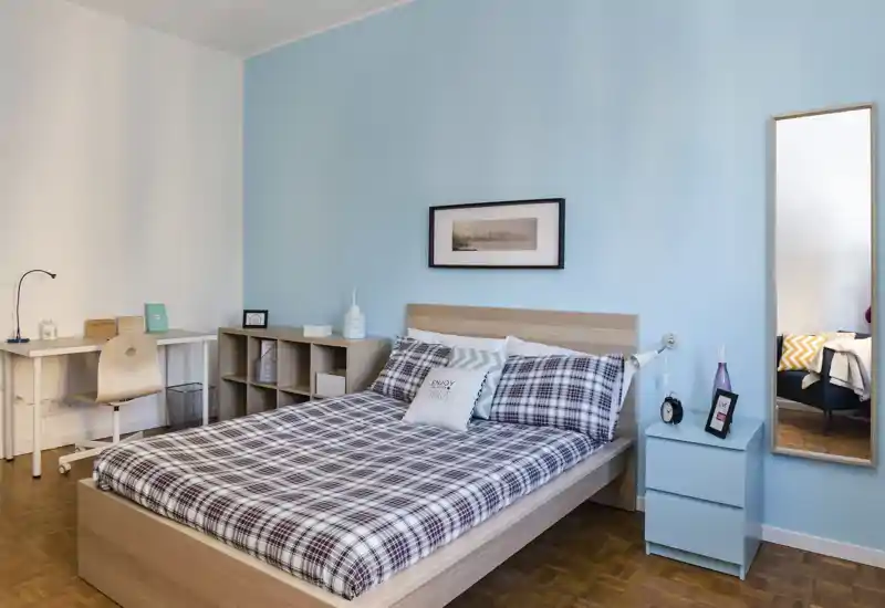 Chambre moderne et lumineuse avec un lit double en linge à carreaux, une table de chevet et un miroir pleine longueur ; bureau simple et étagères latérales pour un espace net et fonctionnel.