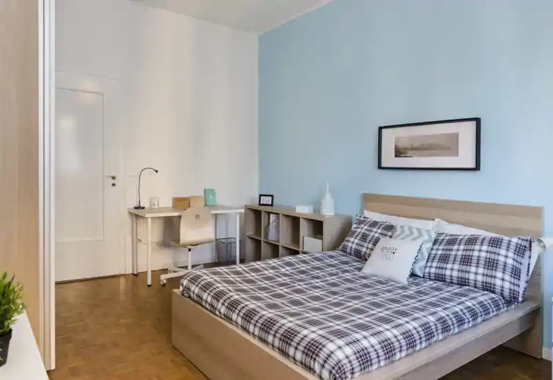 Chambre lumineuse et soignée avec un lit double, couette à carreaux, tête de lit et un espace bureau. Mur bleu pâle et étagères épurées pour une ambiance moderne et apaisante.