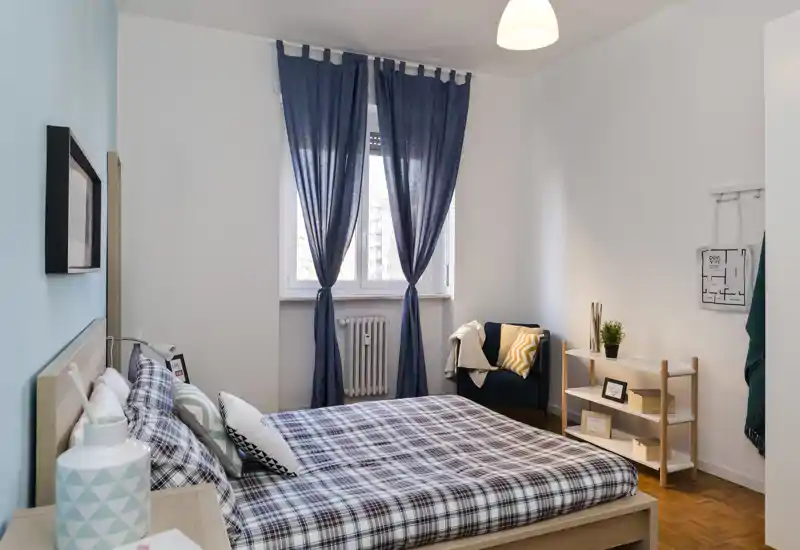 Chambre claire et soignée avec un lit double en linge à carreaux, une fenêtre aux rideaux bleu marine et un fauteuil cosy près d'une petite étagère.