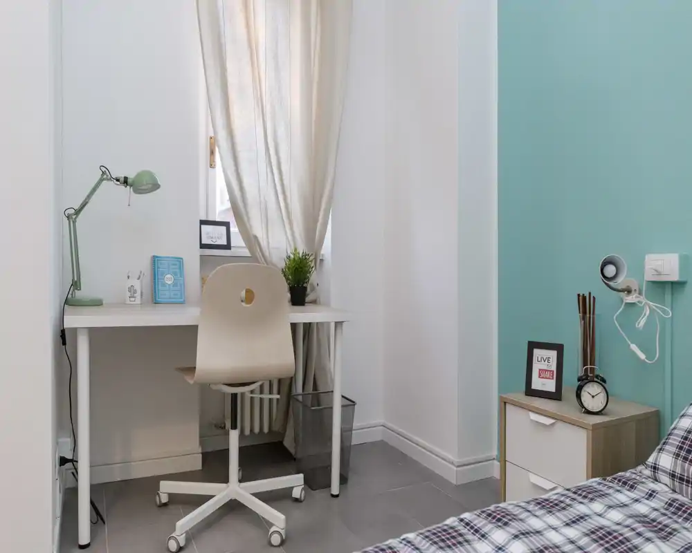 Bright, minimally styled bedroom corner with a desk by the window, a swivel chair, bedside table and part of a plaid bed in the foreground — modern, tidy and well-lit.