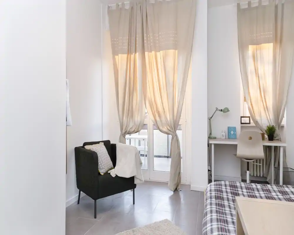 Bright, modern bedroom corner with a comfy bed visible in the foreground, a cozy armchair by the window and a small desk — ideal for a peaceful stay with natural light.