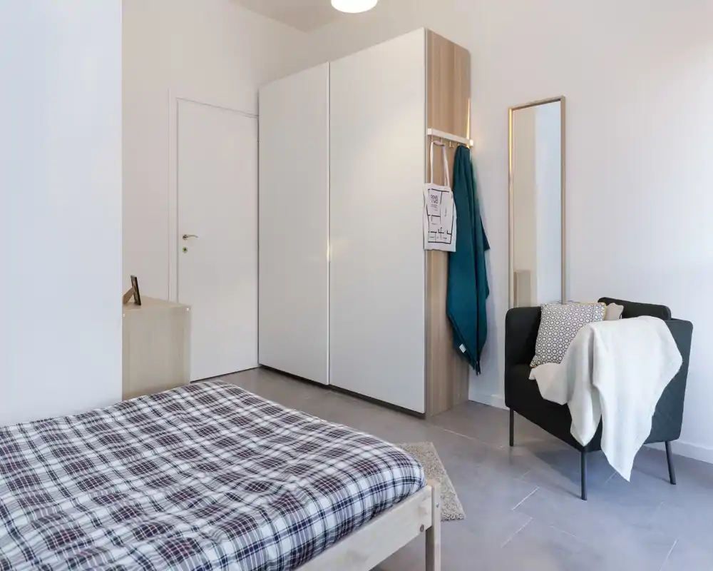 Bright modern bedroom with a plaid-covered wooden bed in the foreground, a large white wardrobe, and a cozy armchair with throws and a full-length mirror — minimal, clean styling ideal for rental photos.