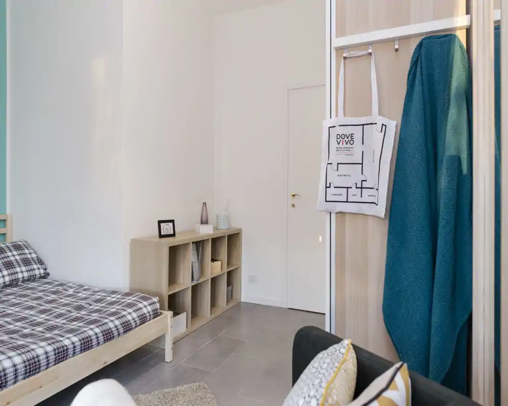 Bright, minimal bedroom with a wooden single bed dressed in checked bedding, low shelving for storage and soft natural light—cozy, practical and neatly styled for rental listings.